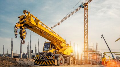 A rugged yellow mobile crane in front of a building construction site with a yellow tower crane in the background and workers on site in hi-visibility jackets