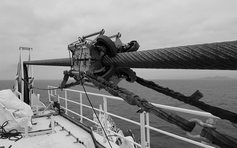 a steel wire rope on a taught line on an offshore vessel with a oil lubricating machine attached to it in black and white